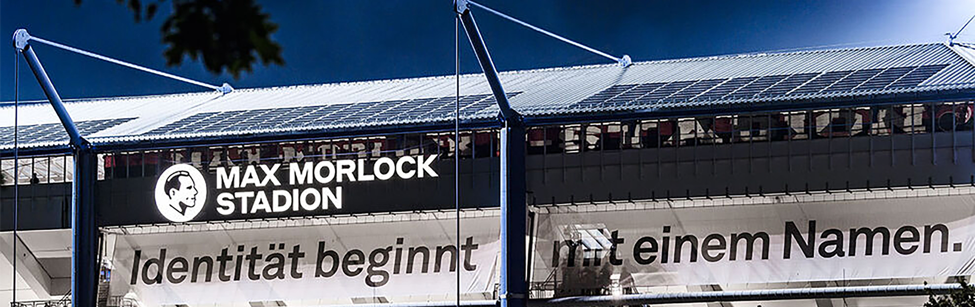 Außenansicht des Max-Morlock-Stadions bei Nacht. Auf der Fassade ist in großen weißen Buchstaben der Schriftzug „MAX MORLOCK STADION“ mit einem stilisierten Porträt daneben zu sehen. Darunter hängt ein Banner mit der Aufschrift „Identität beginnt mit einem Namen.“ Auf dem Stadiondach sind Solarpaneele montiert.